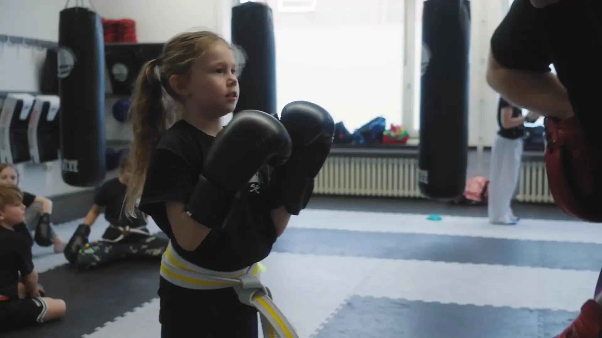 Mädchen mit Boxhandschuhen in Deckung beim Kids-Training, im Hintergrund weitere Kinder und Sandsäcke
