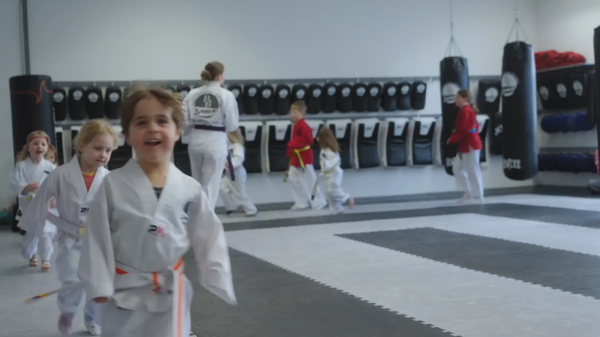 Lachendes Kind im Anzug läuft beim Minis-Training auf die Kamera zu, dahinter weitere Kinder in der Halle